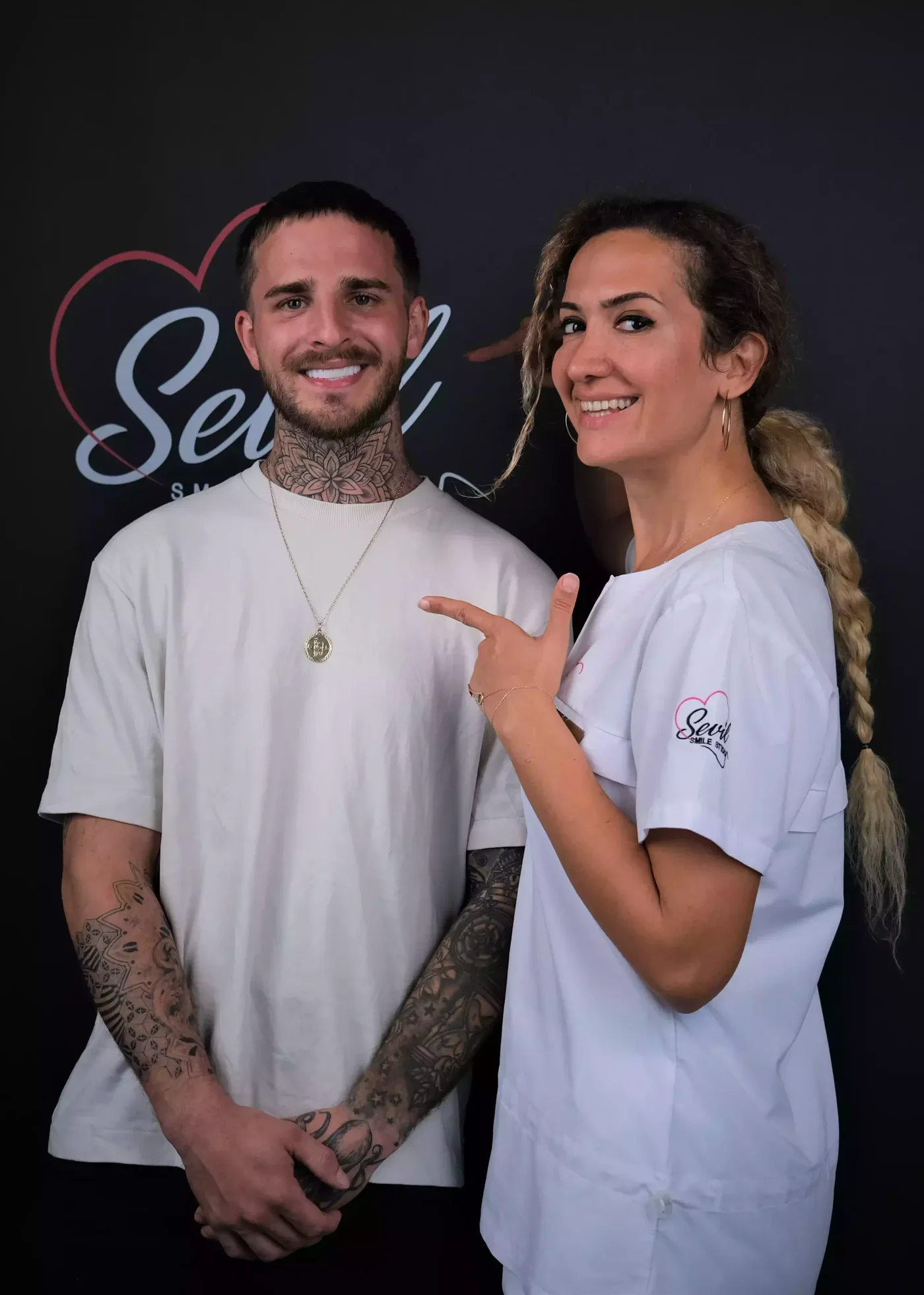 A patient standing beside the clinician who treated her, at the Sevil Smile Studio wall